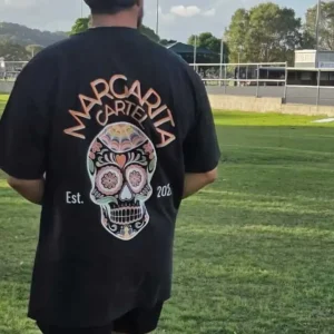 Man wearing a Margarita Cartel black oversized tee with sugar skull design and Est. 2025 branding in a sunny park.